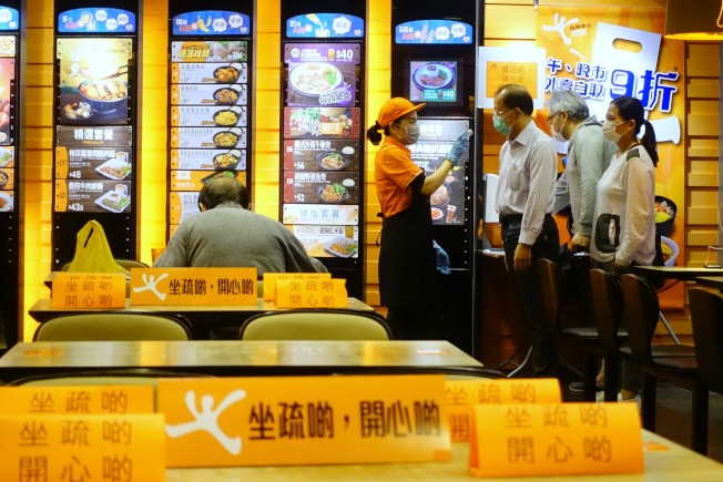 港府規定28日晚上6時開始,所有餐飲業餐廳必須為顧客量體溫,顧客須戴口罩,就餐顧客數目不得超過座位總數一半、每桌最多四人入座等,以減少人員聚集。圖為筲箕灣一家連鎖餐廳店員給顧客量體溫,餐桌上擺放著「坐疏啲,開心啲」(坐疏點,開心點)的提示牌。(中新社)